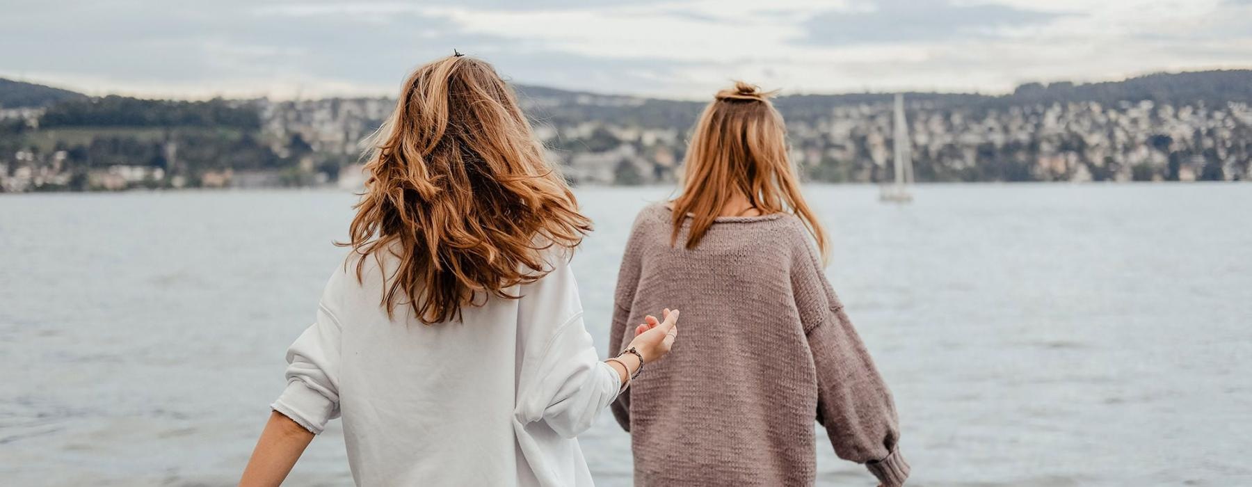 two women walk to the edge of a dock, in a body of water, looking toward the city on the other shore
